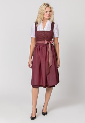Blonde Frau in weißer Bluse und burgunderfarbenem traditionellem Kleid mit Knöpfen und Satinschleife auf der Schürze, trägt schwarze High Heels.