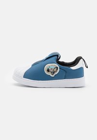 Blue canvas slip-on shoe with white rubber toe cap and sole, featuring a round logo patch on the side. Black interior and pull tab at the heel.
