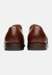 Scarpe in pelle marrone con una finitura liscia, dettagli brogue sui cuciture, punta rotonda e suola bassa in marrone scuro, texturizzata.