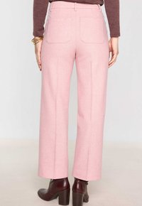 Une personne portant un pantalon large taille haute rose clair, des bottines à talons marron et un haut à manches longues marron, se tenant devant un fond uni.