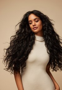 Lang, sort, krøllet hår falder over en lys beige, tætsiddende kjole med høj hals. Baggrunden er i en blød, neutral tone. Guldsmykke som accent.