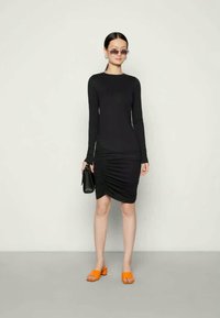 Crna haljina dugih rukava s nabranim detaljima na bokovima, uparena s narančastim natikačama i crnom torbicom. Glatka tekstura, prilagođen dizajn.