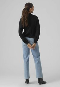 Maglione nero a trama con maniche lunghe, jeans a vita alta azzurri e stivaletti neri. La modella è girata di spalle con le mani incrociate dietro la schiena.