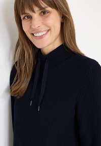 Merinakauhan neulepaita korkealla kauluksella ja kiristysnauhalla. Ominaisuuksiin kuuluu teksturoitu kangas, jossa on ribbireunat, tarjoten mukavan istuvuuden.