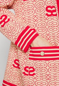 Manche et poche latérale d'un vêtement en maille crème avec des motifs de cœur rouge et de 'S', bordure de manchette et de poche rayée rouge et crème, main à l'intérieur de la poche.