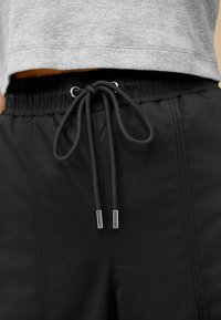 Pantaloni de trening negri cu talie elastică, șnur ajustabil și vârfuri metalice argintii. Prezintă detalii de cusătură laterală pentru un plus de textură.