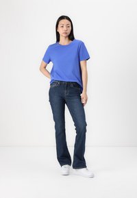 T-shirt en coton bleu à manches courtes, associé à un jean évasé en denim bleu foncé. Le modèle porte des baskets blanches. Tenue simple et décontractée.