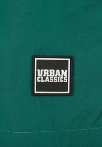 Zöld szövet sima textúrával rendelkezik, amelyen egy téglalap alakú fekete folt található, rajta vastag fehér betűkkel "URBAN CLASSICS" felirattal.