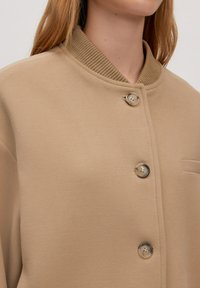 Beige knoopjack met een geribbelde kraag, zachte stofstructuur en een enkele borstzak voor extra detail.
