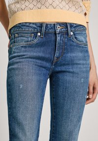 Blå denimjeans med slim fit, blekt utseende, orange sömmar och framfickor. Bärs med en ljusbeige stickad topp.