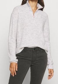 Lichtgrijze, gestructureerde trui met een half-rits kraag en gebreide accenten. Gecombineerd met donkergrijze slim-fit jeans.