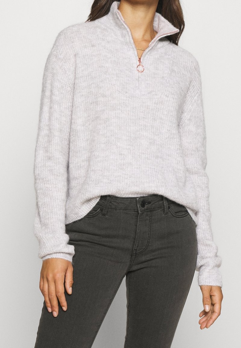 Lichtgrijze, gestructureerde trui met een half-rits kraag en gebreide accenten. Gecombineerd met donkergrijze slim-fit jeans.