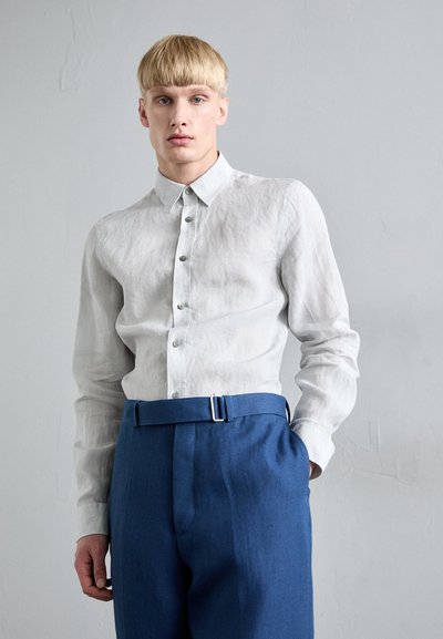 Camisa de lino gris claro con cuello abotonado y botones plateados, combinada con pantalones azules que tienen un cinturón con hebilla de metal.