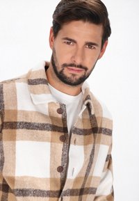 Brun och beige rutig flanellskjorta med träknappar, krage och en mjuk textur. Bärs över en vit t-shirt.