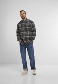 Geruit overhemd in donker groen, rood en wit, gecombineerd met opgerolde blauwe spijkerbroek en lichte sneakers. Het overhemd heeft een button-down kraag.