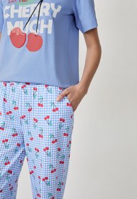 T-shirt in cotone di colore azzurro chiaro con il testo 'CHERRY MUCH' e una grafica di ciliegie, abbinato a pantaloni a quadretti blu con stampa di ciliegie rosse e tasche.