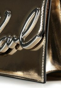 Borsa in oro metallizzato con finitura lucida, caratterizzata da un logo in rilievo con script argentato. I bordi sono cuciti in nero a contrasto.