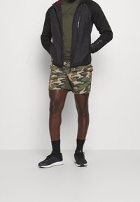 Black lightweight jacket with a zip, olive green top, camouflage shorts, black socks, and running shoes with white sole.