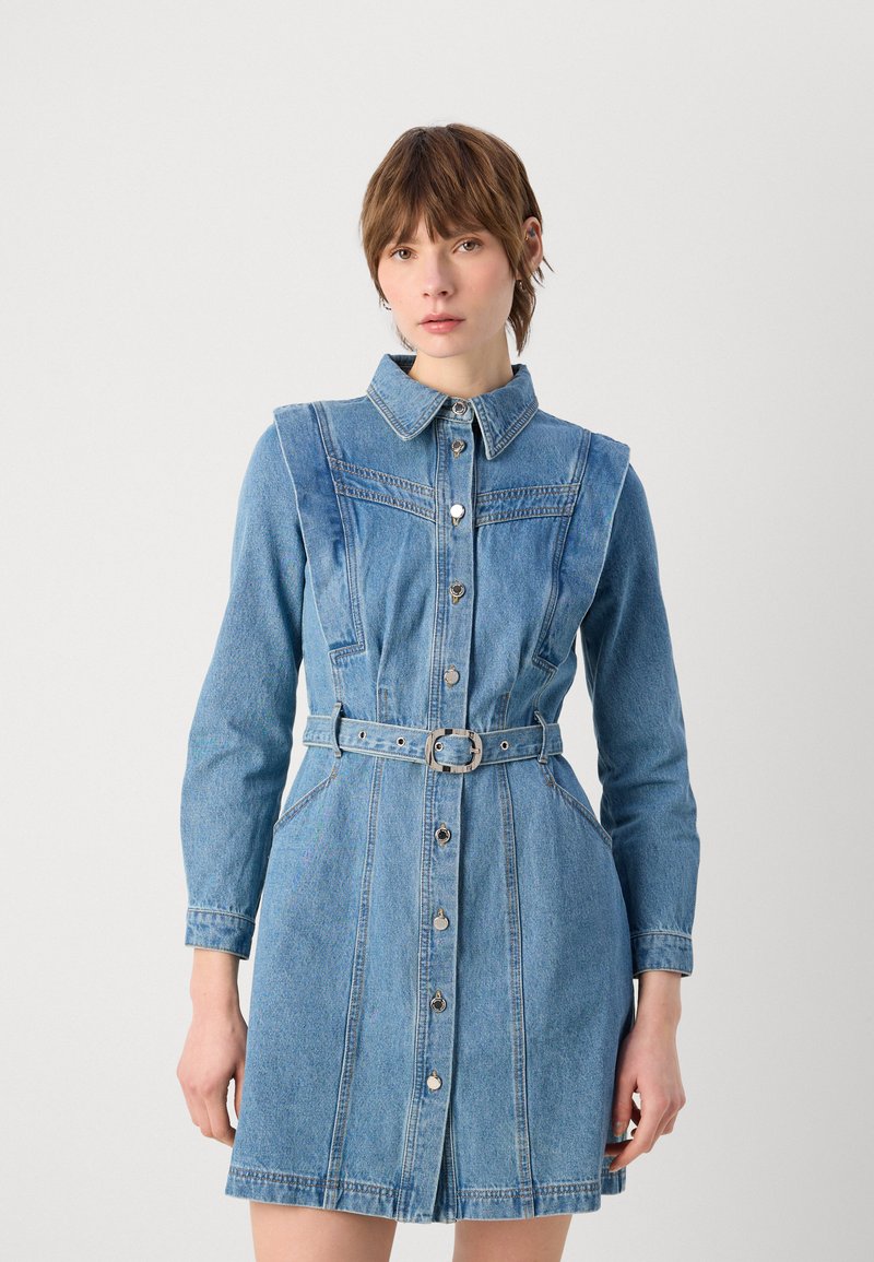 Femme portant une robe en jean bleue boutonnée et ceinturée, à manches longues et col, debout devant un fond clair uni.