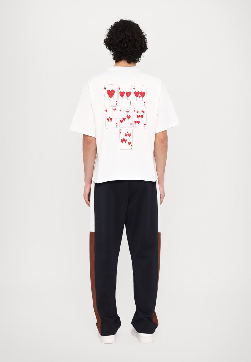 Tricou alb cu un grafic care reprezintă cărți de joc cu inimă roșie pe spate. Asortat cu pantaloni bleumarin cu panouri laterale maro și albe.
