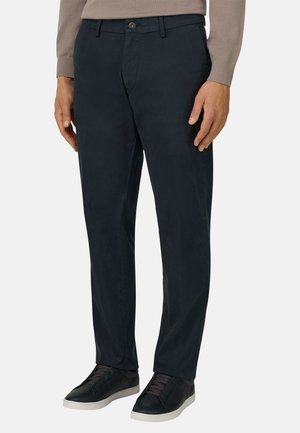 Dunkelblaue Chino-Hosen für Herren mit Gürtelschlaufen, vorderer Knopf- und Reißverschluss, kombiniert mit schwarzen Low-Top-Sneakers und einem beigefarbenen Langarmshirt.