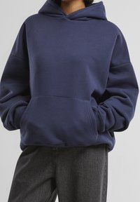 Sweat à capuche bleu marine avec une grande poche avant, coupe oversize, poignets côtelés et capuche à cordon, fabriqué dans un tissu doux. Porté avec un jean foncé.