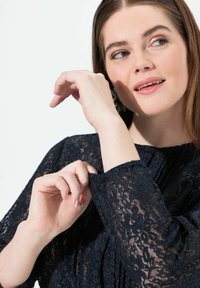 Top in pizzo blu navy con maniche lunghe, caratterizzato da motivi floreali e polsini arricciati. Texture liscia con una finitura semi-trasparente.