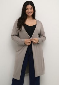 Cardigan lungo e aperto davanti in beige chiaro, realizzato in tessuto morbido. Presenta maniche lunghe e una vestibilità comoda, abbinato a una maglietta nera e jeans scuri.