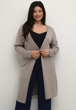 Lange, open voorkant cardigan in lichtbeige, gemaakt van zachte stof. Heeft lange mouwen en een relaxed fit, gecombineerd met een zwarte top en donkere jeans.