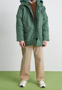 Grüne Steppjacke mit Kapuze, zwei vorderen Taschen und Knopfverschluss. Kombiniert mit beigen Hosen und weißen Turnschuhen auf grünem Boden.