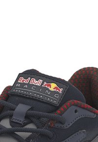 Gros plan sur la languette sombre d'une chaussure de sport avec le logo de l'équipe Red Bull Racing de Formule 1 et une doublure intérieure à carreaux noirs et rouges.