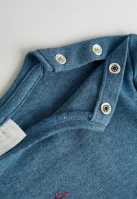 Col enbody pour bébé bleu avec trois boutons-pression argentés et détails de couture, montrant une partie d'une étiquette de vêtement blanche.