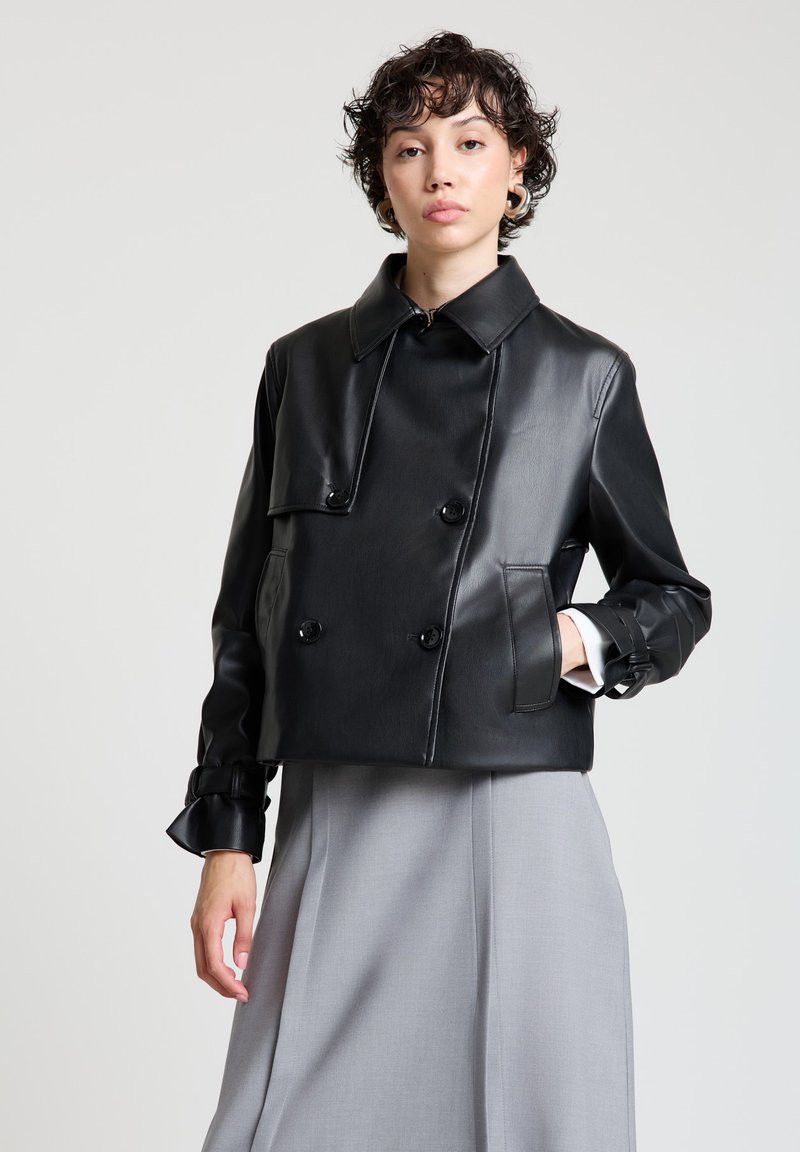 Schwarze, kurze Lederjacke mit doppelreihigen Knöpfen, großem Kragen, Seitentaschen und gerafften Manschetten, kombiniert mit einem grauen Rock.