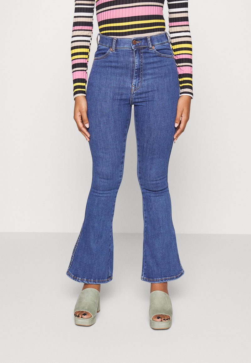 Jeans flare de cintura alta em denim azul médio com uma textura suave e uma bainha ligeiramente desgastada, combinados com sandálias plataforma cinzas.