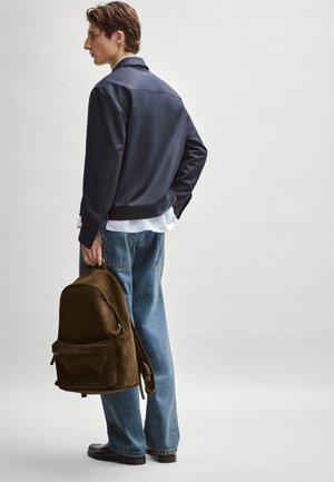 Mladý muž v tmavé bundě, modrých džínách a černých botách drží hnědý batoh na jednobarevném pozadí.