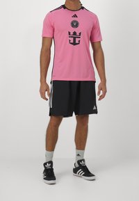 Mies, joka seisoo päällään vaaleanpunainen Inter Miami CF -jalkapallopaita, mustat Adidas-shortsit, valkoiset Adidas-sukat ja mustat Adidas-tossut.