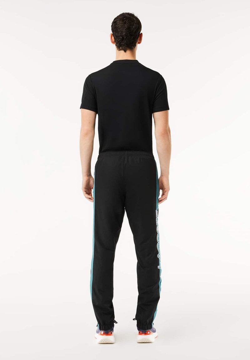 Tenue de sport noire, comprenant un t-shirt à manches courtes et un pantalon ajusté avec des rayures latérales bleues et un logo blanc. Tissu lisse, ceinture élastique.