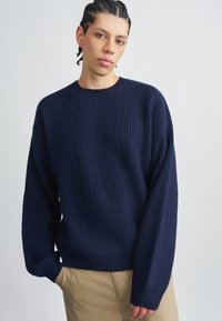 Maglione lavorato a maglia blu navy con toppe testurizzate, polsini e orlo a coste. Presenta un collo rotondo e una vestibilità comoda. Indossato sopra pantaloni beige.