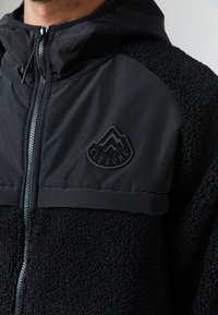Gros plan sur une personne portant une veste polaire noire à fermeture éclair avec une capuche et un patch logo montagne étiqueté "Closure" sur la poitrine.