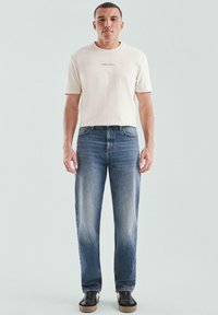 Mężczyzna stojący przodem, ubrany w kremowy t-shirt z małym napisem, niebieskie dżinsy i czarne tenisówki z beżowymi podeszwami.