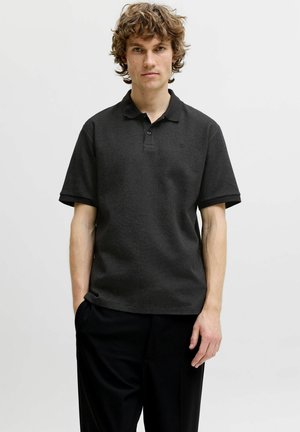 Giovane uomo con capelli ricci che indossa una polo nera a maniche corte con trama e pantaloni neri, in piedi davanti a uno sfondo chiaro e uniforme.