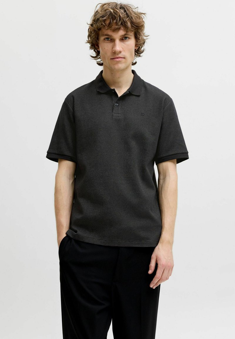 Jeune homme aux cheveux bouclés portant un polo noir texturé à manches courtes et un pantalon noir, debout devant un fond clair uni.