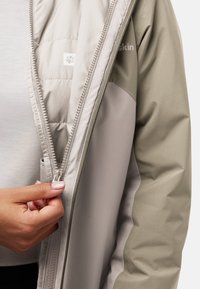 Lichte, bicolor jas in beige en olijfgroen, voorzien van een rits aan de voorkant en gewatteerde voering. Glad textuur met subtiele branding.