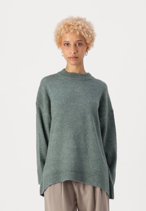 Übergroßer grüner Pullover aus weichem, strukturiertem Stoff mit Rundhalsausschnitt und geripptem Saum. Kombiniert mit lockeren beigen Hosen.