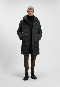 Abrigo de plumas negro con un cuello alto, botones de presión y capucha. Combinado con un suéter de cuello alto negro y pantalones marrones, además de botas negras con cordones.