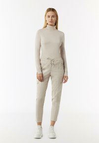 Lichtbeige coltrui gecombineerd met lichtbeige joggingsbroek met elastische tailleband, zijritsen en cargozakken. Witte sneakers.