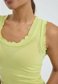 Mujer vistiendo una camiseta sin mangas de canalé verde claro con bordes con volantes, mostrada desde el cuello hasta la cintura contra un fondo liso.