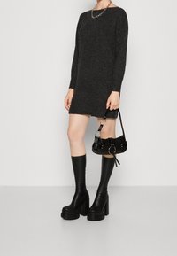 Modelka v těsně přiléhajících tmavých svetrových šatech s dlouhými rukávy, v černých vysokých platformových botách po kolena a s malou černou kabelkou.