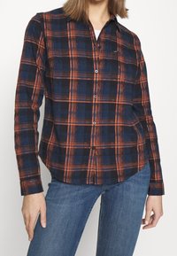 Camicia button-up in fantasia scozzese blu marina e arancione, con maniche lunghe e orlo curvo. Realizzata in un tessuto morbido e leggero.
