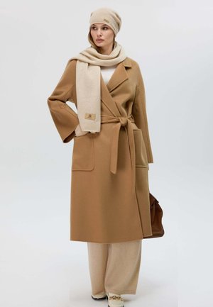 Beiger Wollmantel mit Revers, tailliertem Gürtel und zwei Vordertaschen. Kombiniert mit einem hellbeigen Schal und Hut. Das Model hält eine braune Tasche.